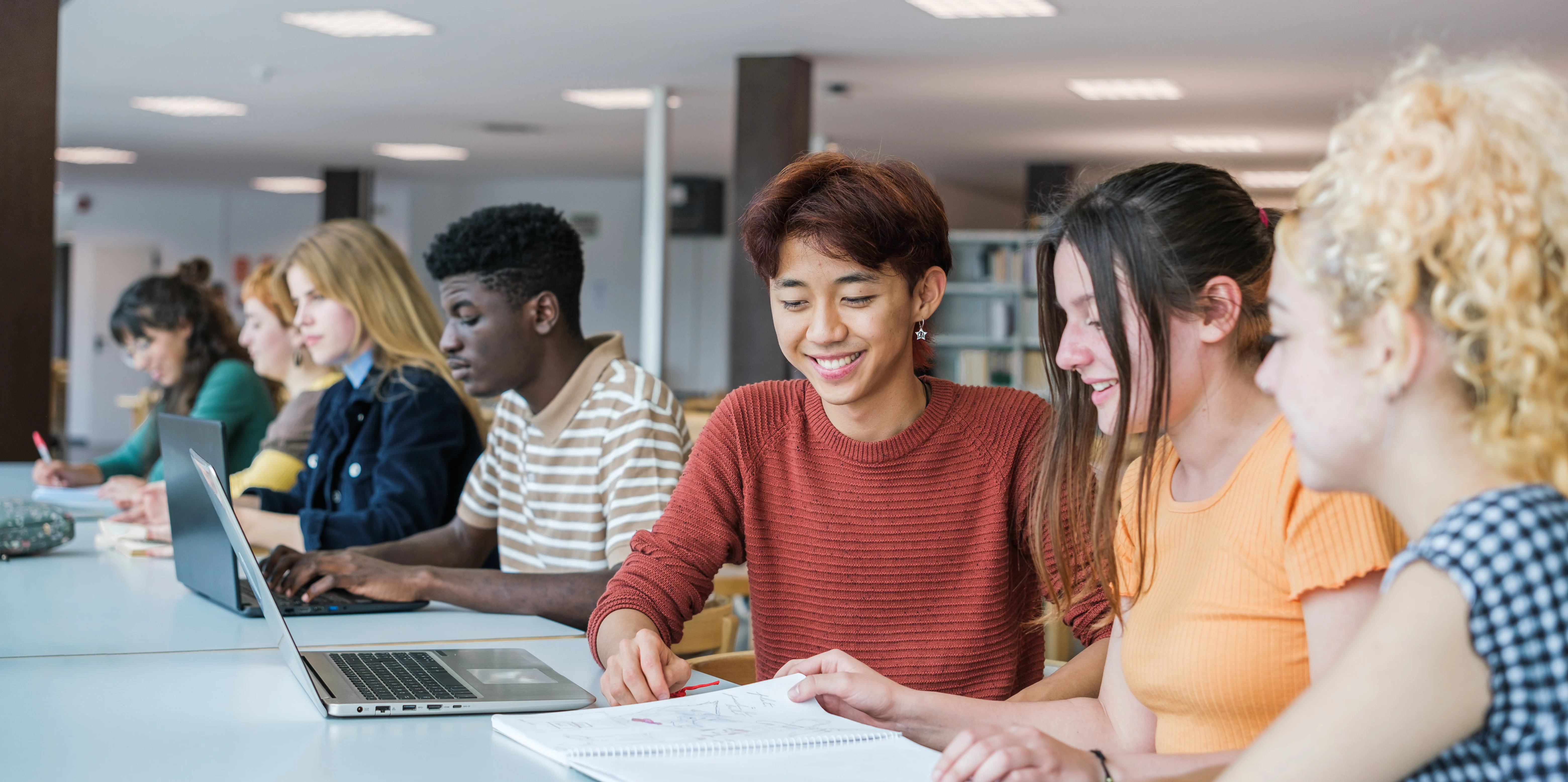 Estudantes compartilham conhecimentos e utilizam notebooks em um ambiente de estudo colaborativo, destacando o papel da tecnologia na educação e no desenvolvimento de novas formas de aprendizagem.