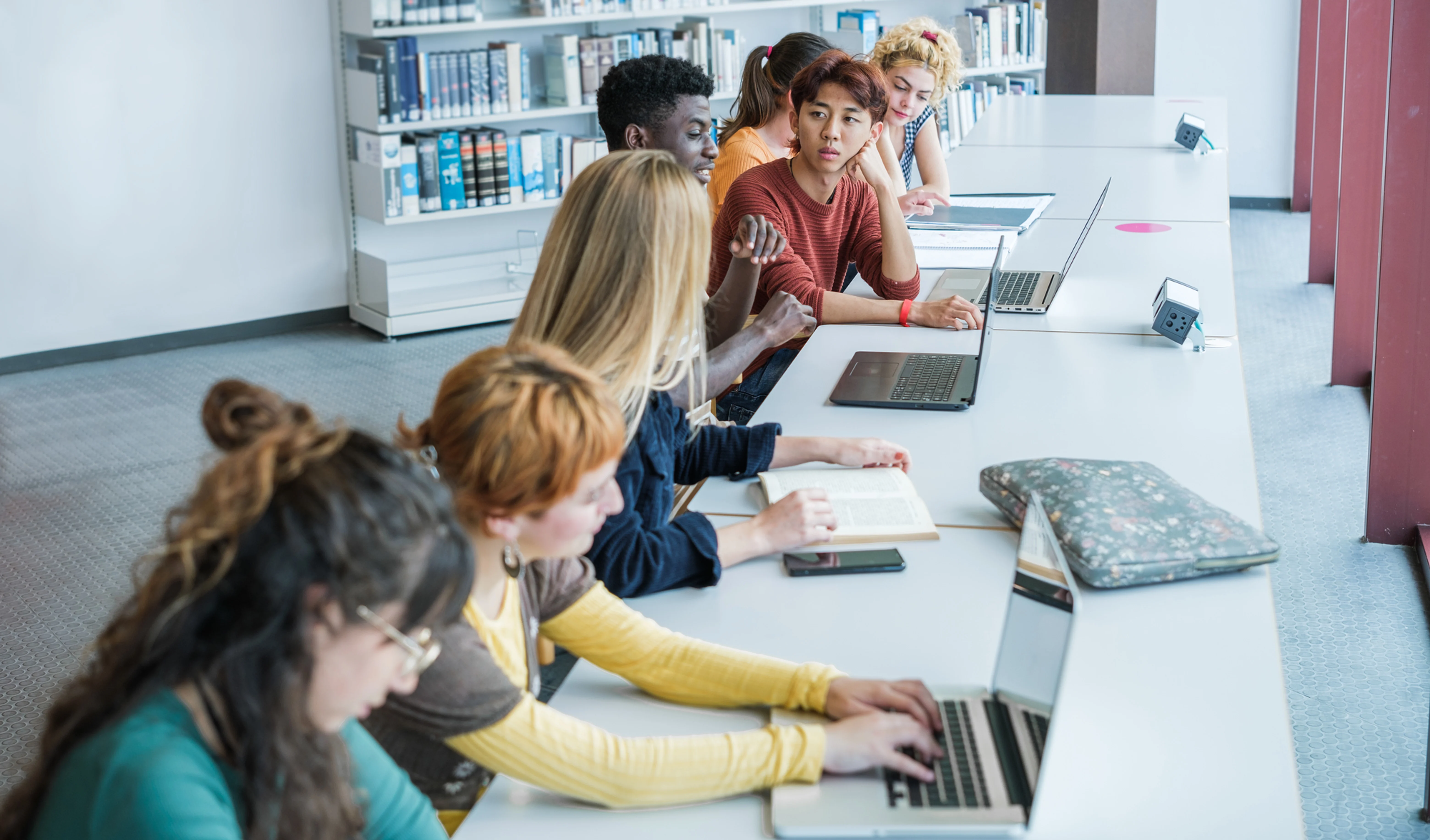 Estudantes utilizam notebooks e materiais de estudo em uma biblioteca, ilustrando a integração das tecnologias digitais ao ambiente educacional e as novas possibilidades de aprendizagem colaborativa.