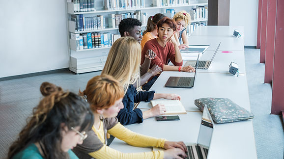 Estudantes utilizam notebooks e materiais de estudo em uma biblioteca, ilustrando a integração das tecnologias digitais ao ambiente educacional e as novas possibilidades de aprendizagem colaborativa.