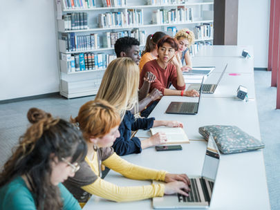 Estudantes utilizam notebooks e materiais de estudo em uma biblioteca, ilustrando a integração das tecnologias digitais ao ambiente educacional e as novas possibilidades de aprendizagem colaborativa.